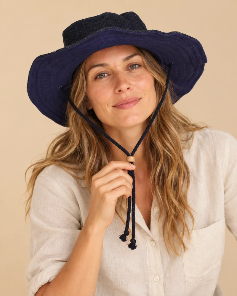 Female model wearing Navy Blue KTM Safari Hat with Chin String and natural hemp shirt, holding the adjustable chin cord against a beige background.