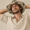 Ellie wearing Large Leaf Print Hemp Hat and natural hemp shirt, posing with his hands behind his head against a beige background.