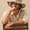 Male model Ellie sitting on a wooden chair wearing Large Mushroom Print Hemp Hat, natural hemp shirt and wide brim with brown mushroom illustrations.