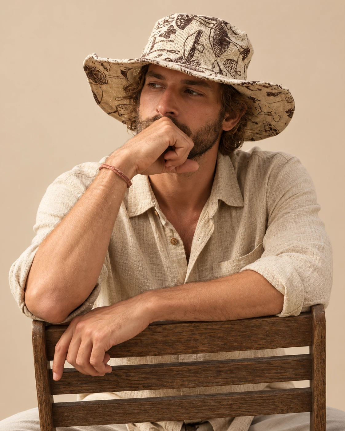 Male model Ellie sitting on a wooden chair wearing Large Mushroom Print Hemp Hat, natural hemp shirt and wide brim with brown mushroom illustrations.