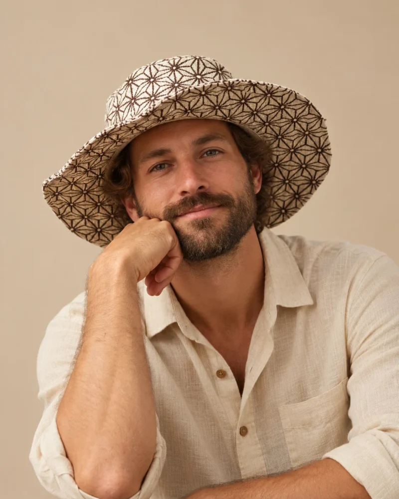 male model Maya wearing a beige wide-brim Himalayan hemp hat with brown cobweb print and natural linen shirt against a beige studio background.
