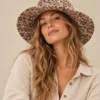 Female model Maya wearing a beige wide-brim Himalayan hemp hat with brown floral print and natural linen shirt against a beige studio background.